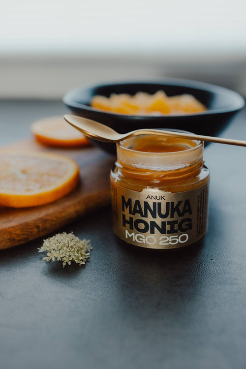 ANUK Manuka Honig MGO250 im Glas, geöffnet, goldener Honig sichtbar, mit Löffel und Obst eindekoriert