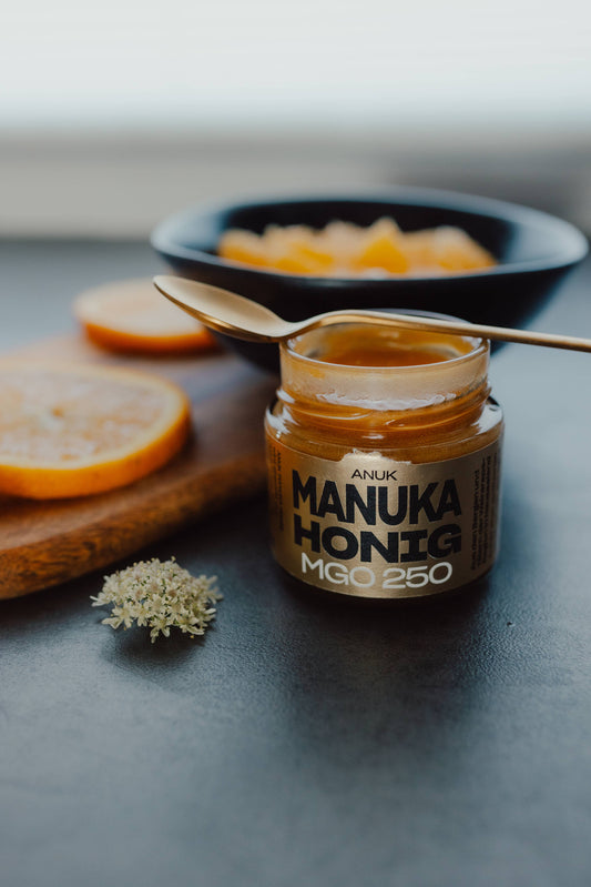 ANUK Manuka Honig MGO250 im Glas, geöffnet, goldener Honig sichtbar, mit Löffel und Obst eindekoriert 