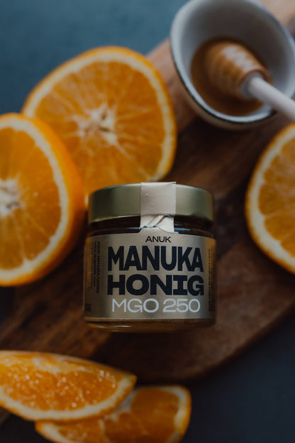 Manuka Honig 250 MGO von Oben neben Orangen auf Holzbrett, Honiglöffel mit Honig daneben
